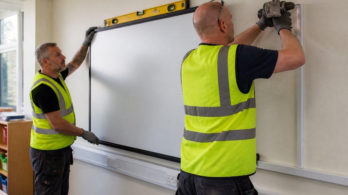 Two installation technicians in safety gear mounting a large interactive display on a classroom wall, using a spirit level and drill, with cables neatly routed in trunking